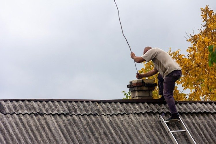 chimney-sweep-man-cleans-chimney-from-soot-roof-village-house-against-gray-sky-with-copy-space-preparing-heating-season